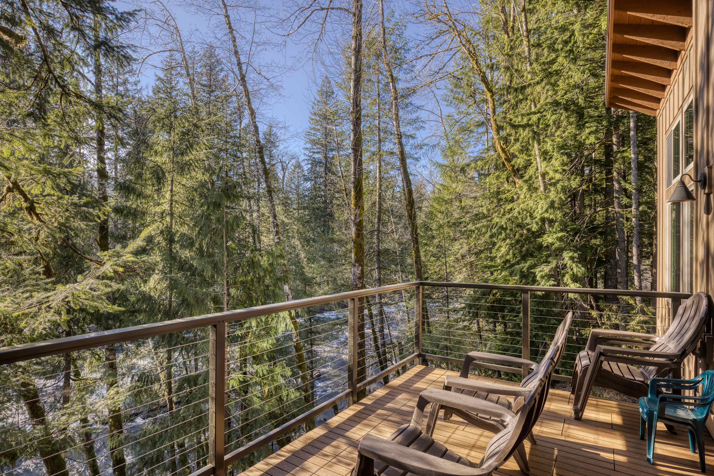 A spacious wooden balcony with modern cable railings offers panoramic views of a lush green forest and a winding river below, furnished with several dark outdoor chairs.