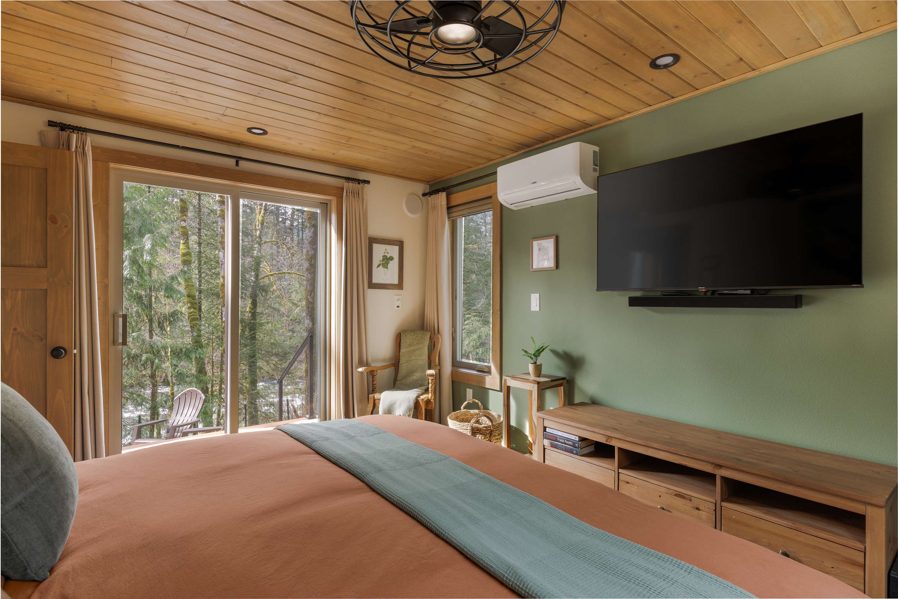 A bedroom with a green accent wall, a mounted TV, and a wooden plank ceiling. A large bed with orange bedding faces a sliding glass door that opens to a deck with views of a lush forest.
