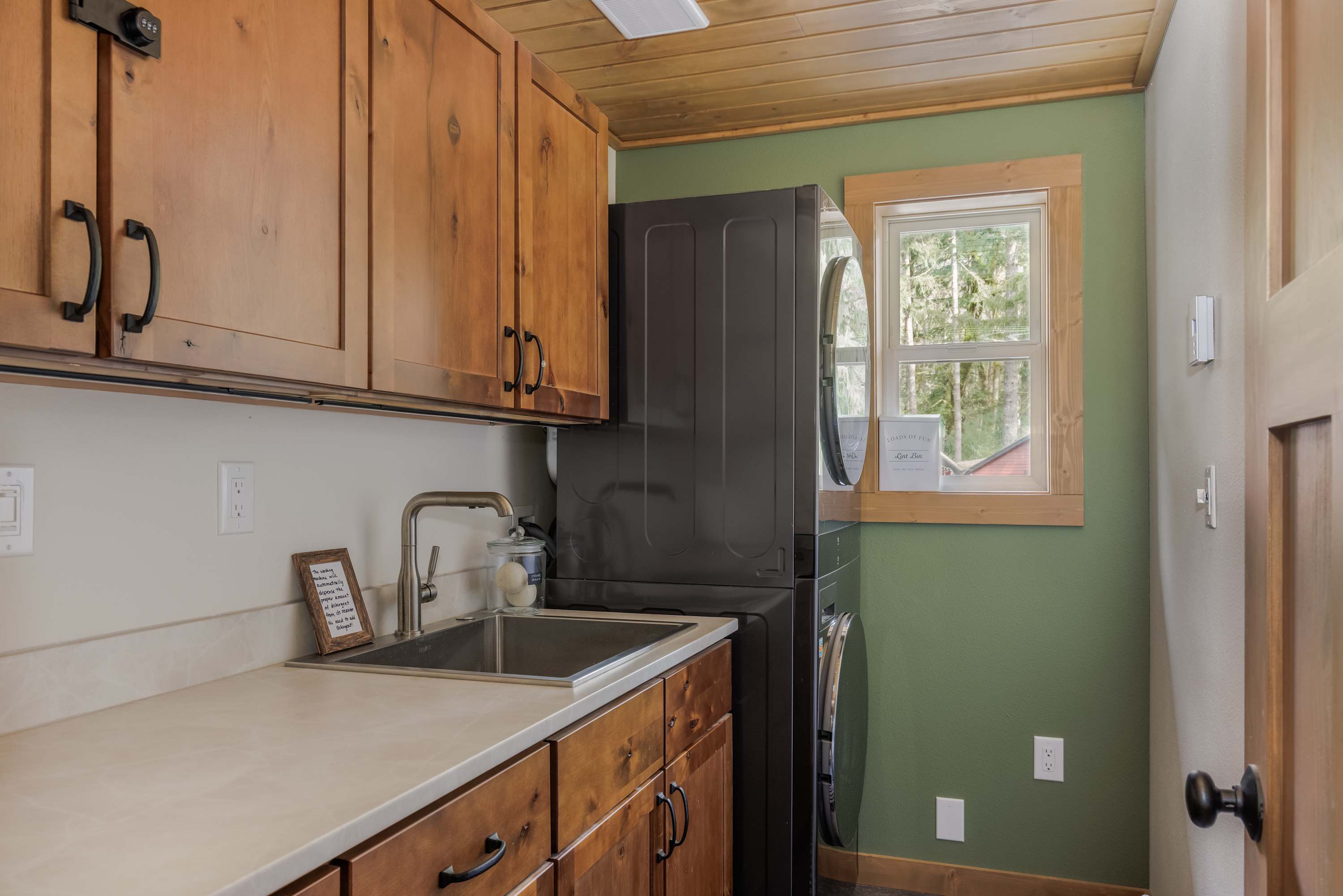 A functional laundry room features a stacked black washer and dryer unit next to a window, with a utility sink set into a light-colored countertop. Wooden cabinets provide storage above and below, and the room has a warm wooden plank ceiling and green accent wall.