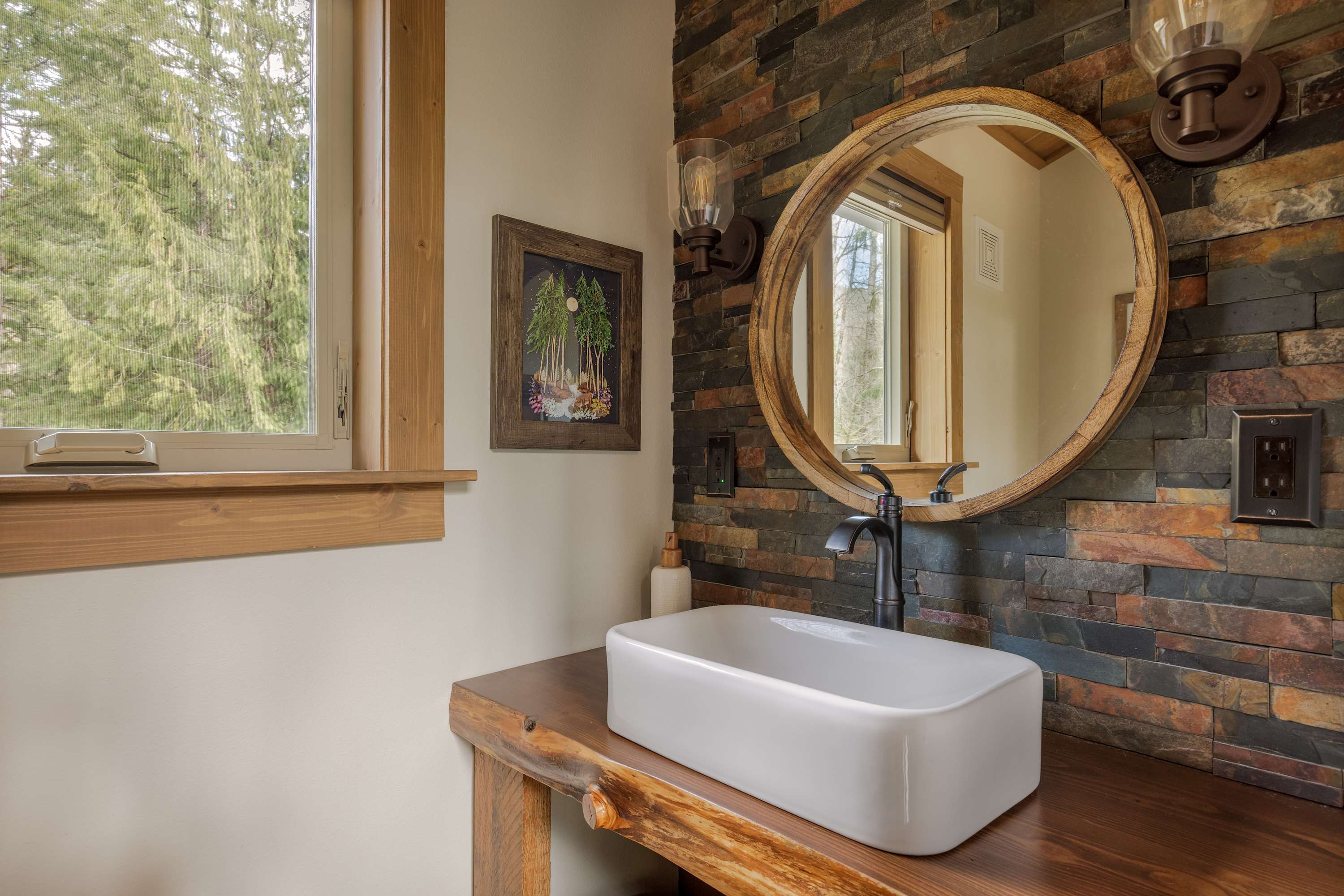 Rustic bathroom featuring a vessel sink on a live-edge wooden vanity, with a round mirror and sconce lighting against a stacked stone accent wall. A window offers a glimpse of the outdoors.
