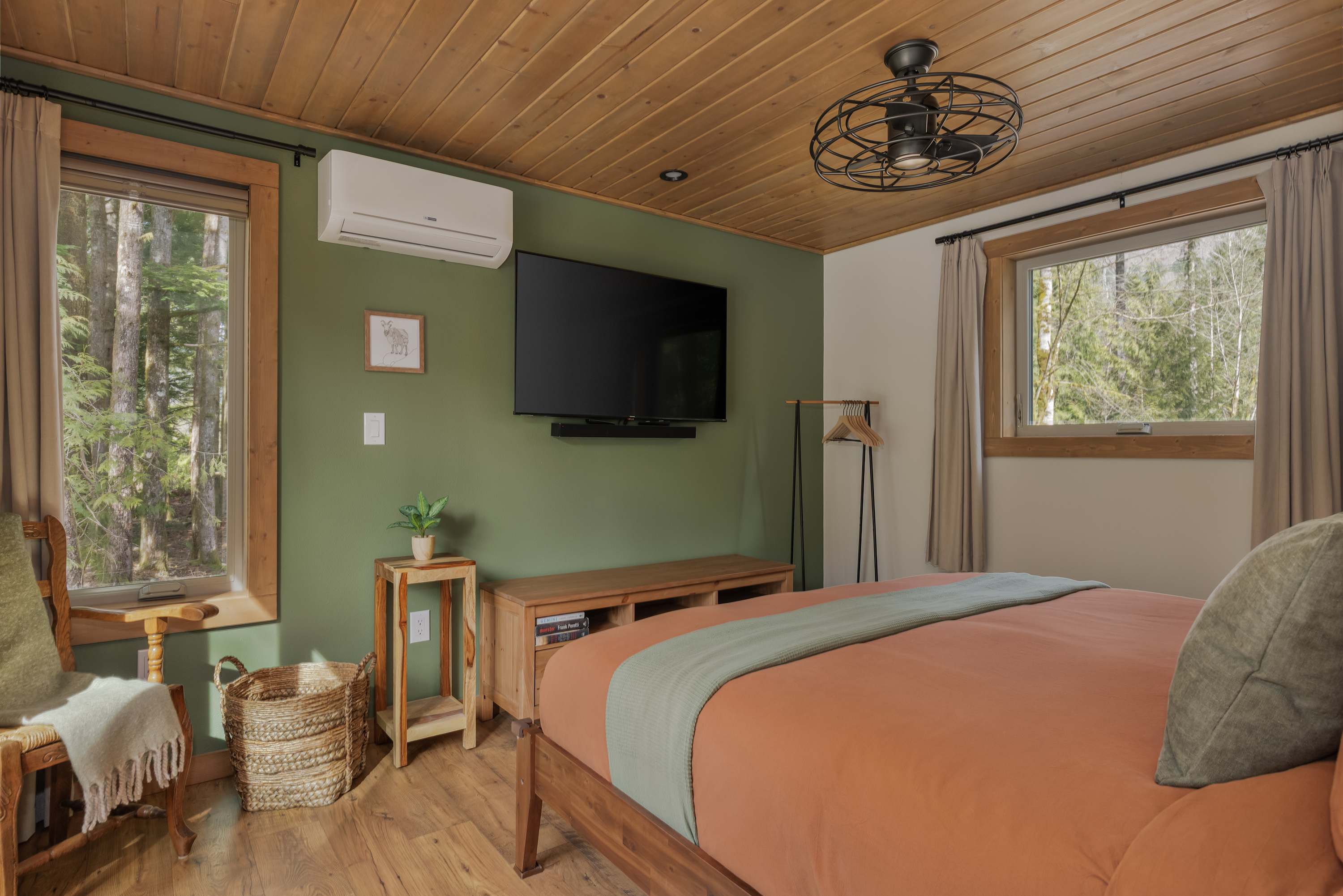 A bedroom with a striking green accent wall featuring a mounted flat-screen TV and an air conditioning unit. The room has a wooden plank ceiling, a bed with orange bedding, and a large window overlooking a wooded area.