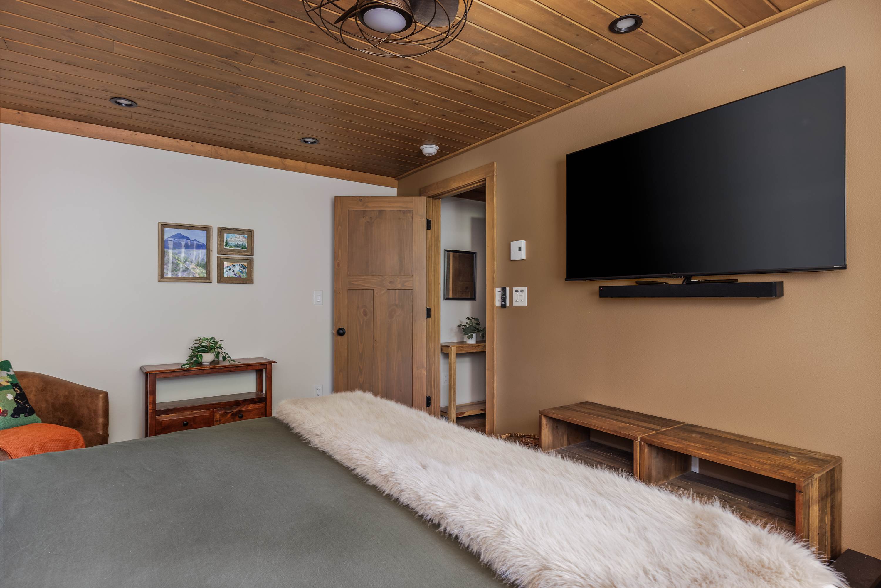 The bedroom features a large wall-mounted flat-screen TV with a soundbar above a low wooden console. A wooden plank ceiling and warm-toned walls create a cozy atmosphere, with a glimpse into an adjacent hallway through an open wooden door.