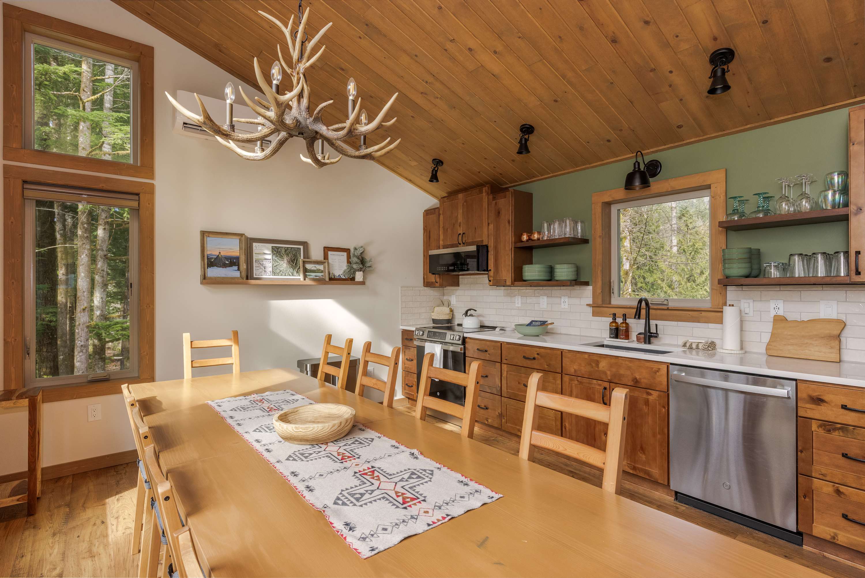 Bright dining room with a long wooden table and chairs, featuring a rustic antler chandelier and a wood-paneled vaulted ceiling. The kitchen with green accent wall and open shelving is visible in the background.