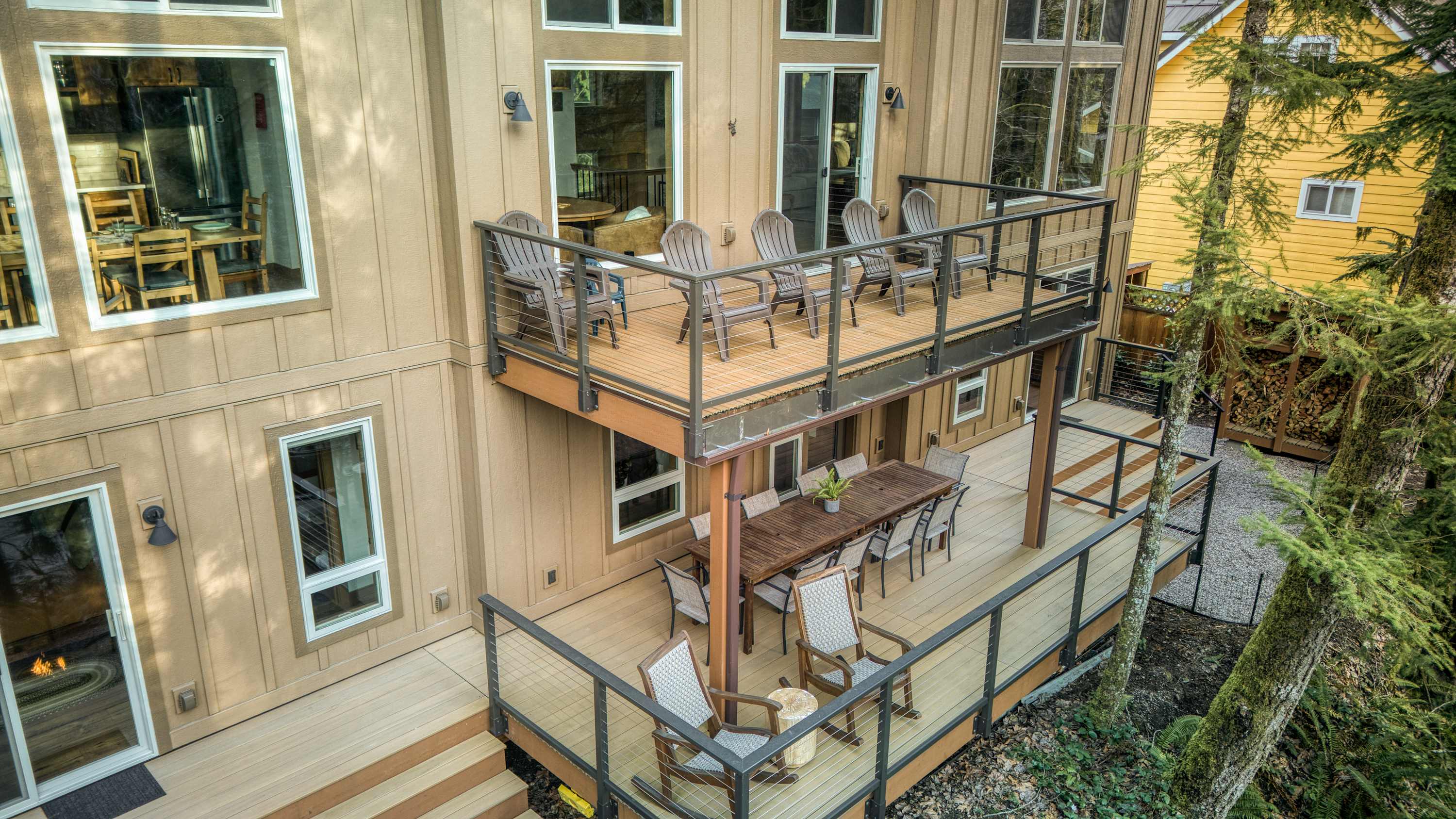An aerial view showcases the back of a multi-story house with light brown siding, featuring two expansive wooden decks equipped with outdoor dining and seating areas, all surrounded by lush trees.