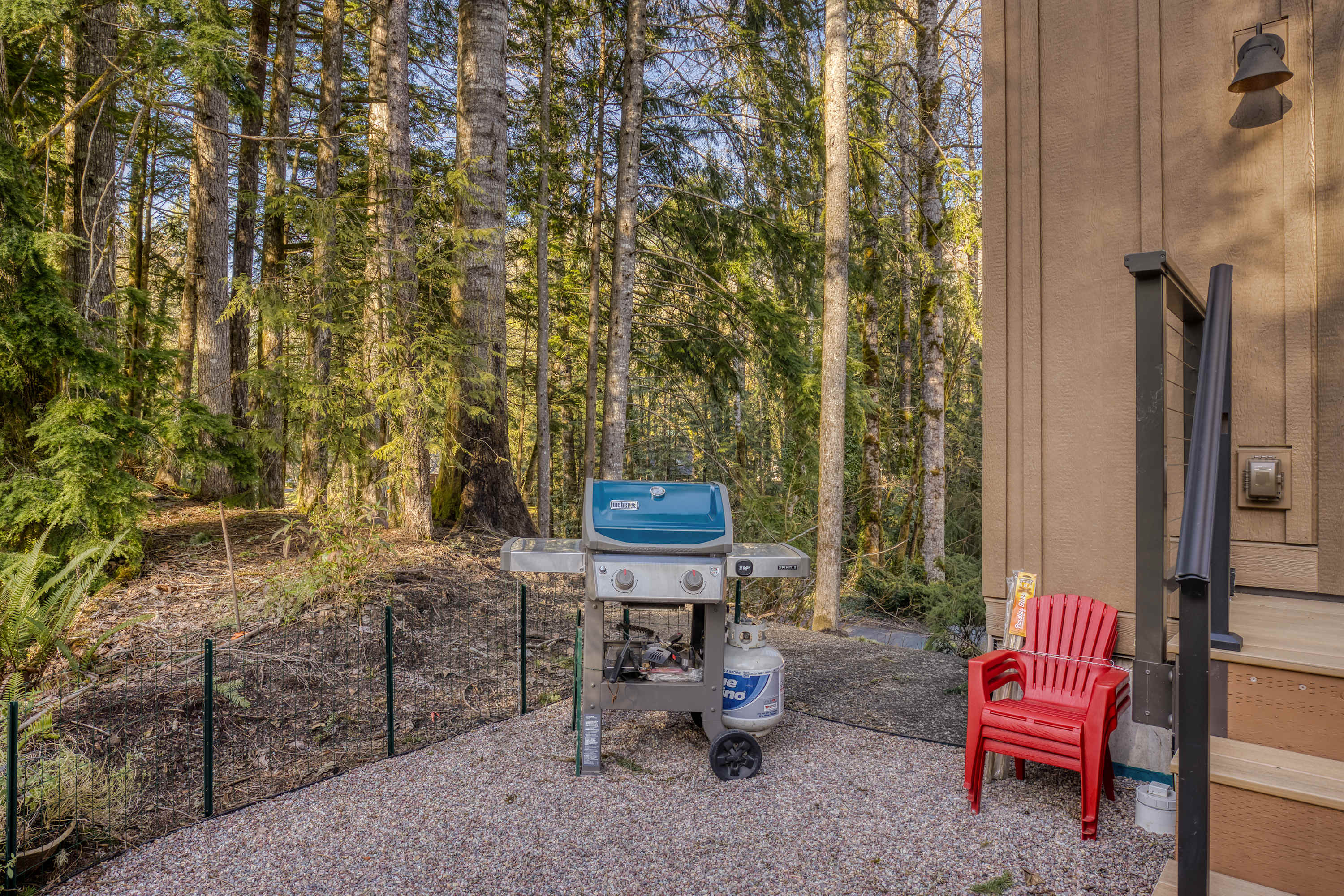 A blue Weber gas grill with a propane tank stands on a gravel patio next to the house, surrounded by a lush forest. A single red plastic chair sits near the wooden deck stairs, offering a casual outdoor cooking and seating spot.