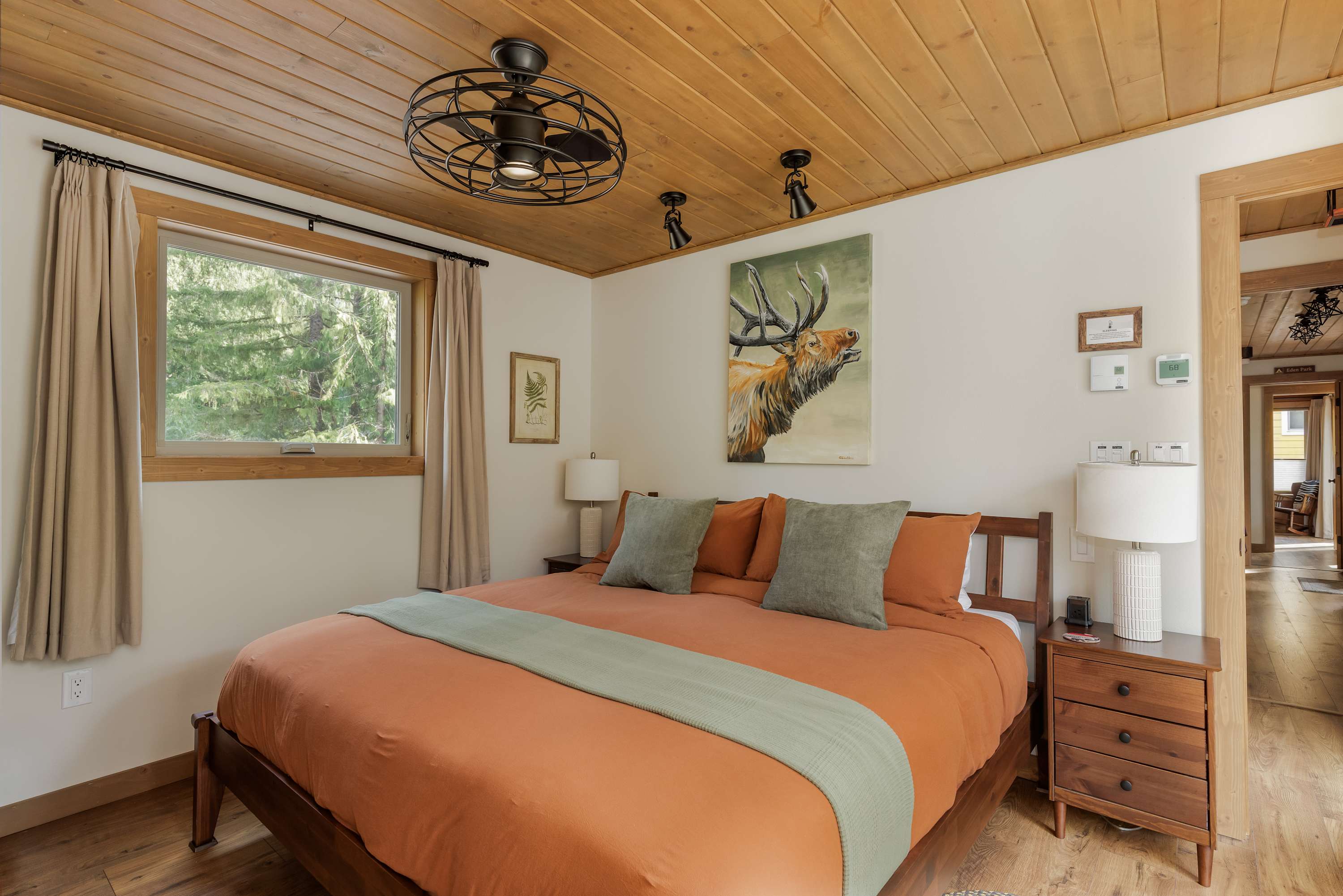 A cozy bedroom with a wooden plank ceiling, a large bed with orange bedding and green pillows, flanked by wooden nightstands with lamps. A large painting of an elk hangs above the bed, and a window offers a view of trees.