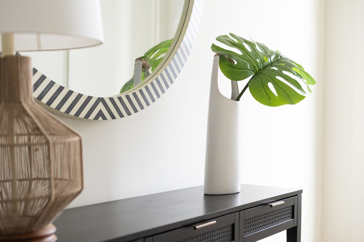 A stylish foyer features a black console table with a woven lamp and a white vase holding a monstera leaf, positioned beneath a round mirror with a black and white geometric pattern. The walls are white, and a light-colored wooden floor is visible.
