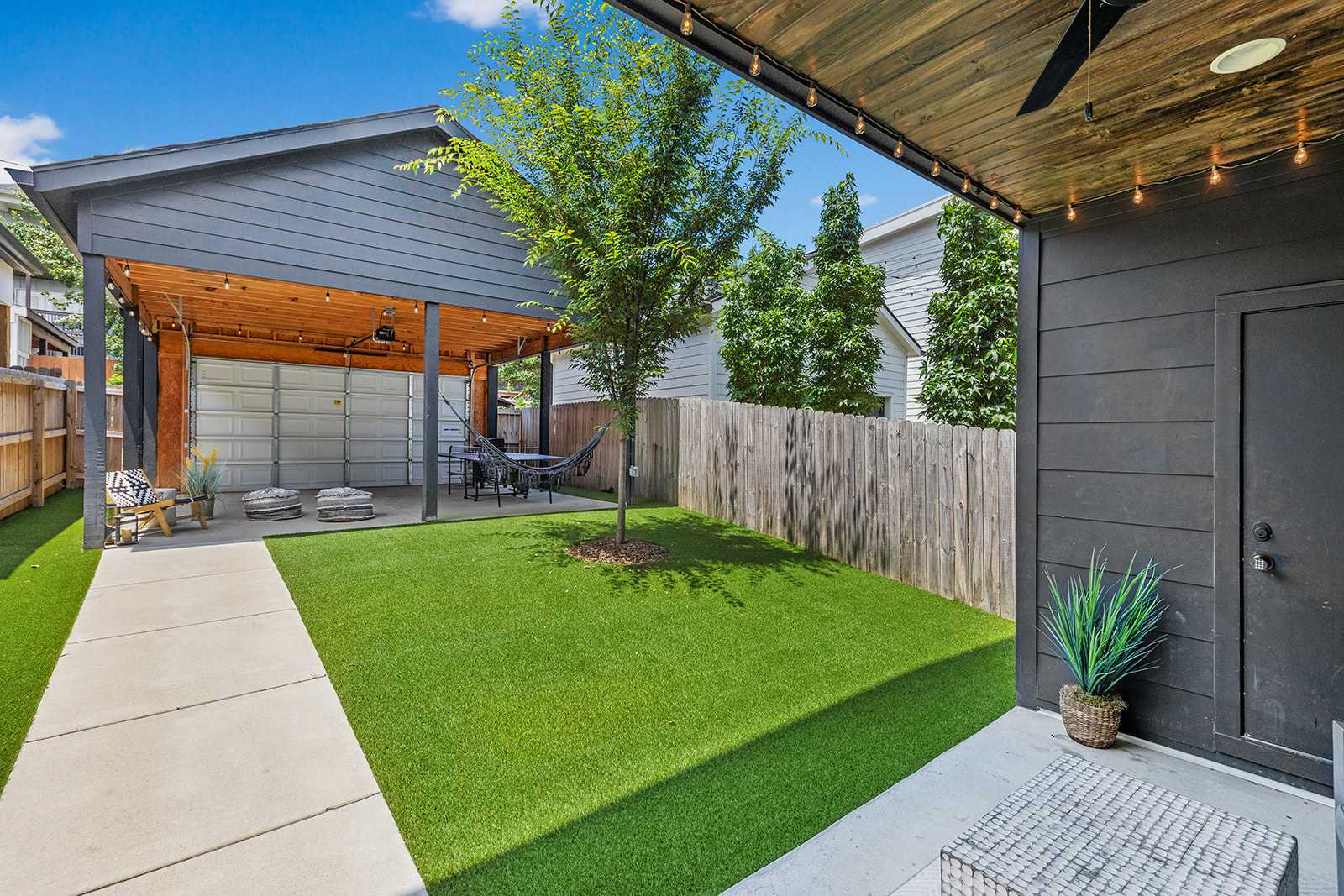 A spacious backyard featuring a covered patio area with string lights and a ceiling fan, a lush green artificial turf lawn with a small tree, and a tall wooden privacy fence. A concrete path leads through the yard.