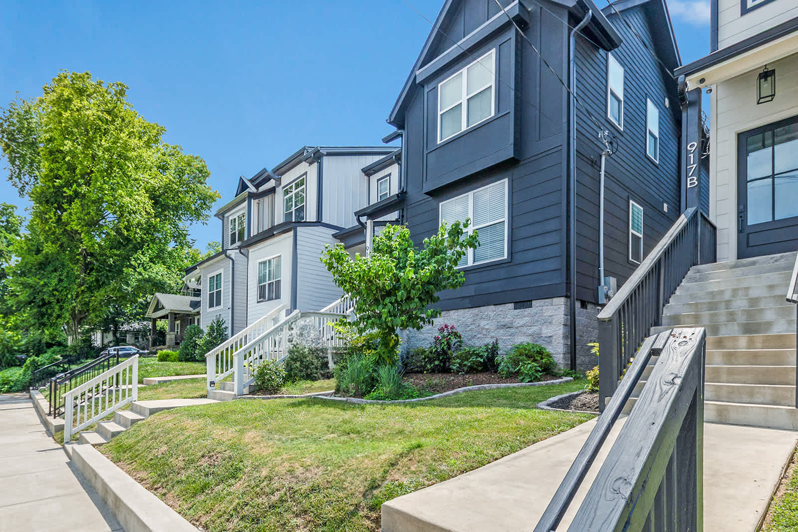 A modern black house with white trim and a white staircase, featuring a small landscaped front yard with a tree, situated in a neighborhood with similar contemporary homes.