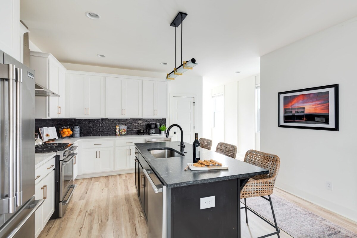 Modern kitchen with a dark island and two woven bar stools, white upper and lower cabinets, a stainless steel refrigerator, and a striking black subway tile backsplash, all on light hardwood floors.