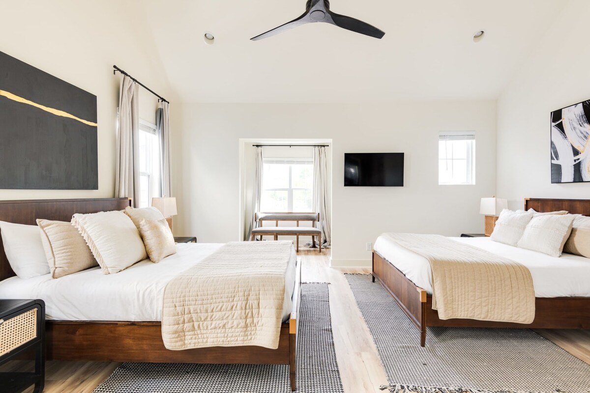 Wide view of a bright bedroom with two wooden beds, each dressed in white and beige, separated by a patterned area rug. Large windows and a modern ceiling fan contribute to the airy feel of the room.