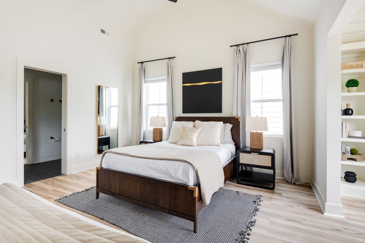 Bright bedroom with a wooden bed frame, white bedding, and beige throw, positioned on a patterned area rug. A built-in bookshelf filled with decor is visible in an alcove, and a doorway leads to an adjacent bathroom.
