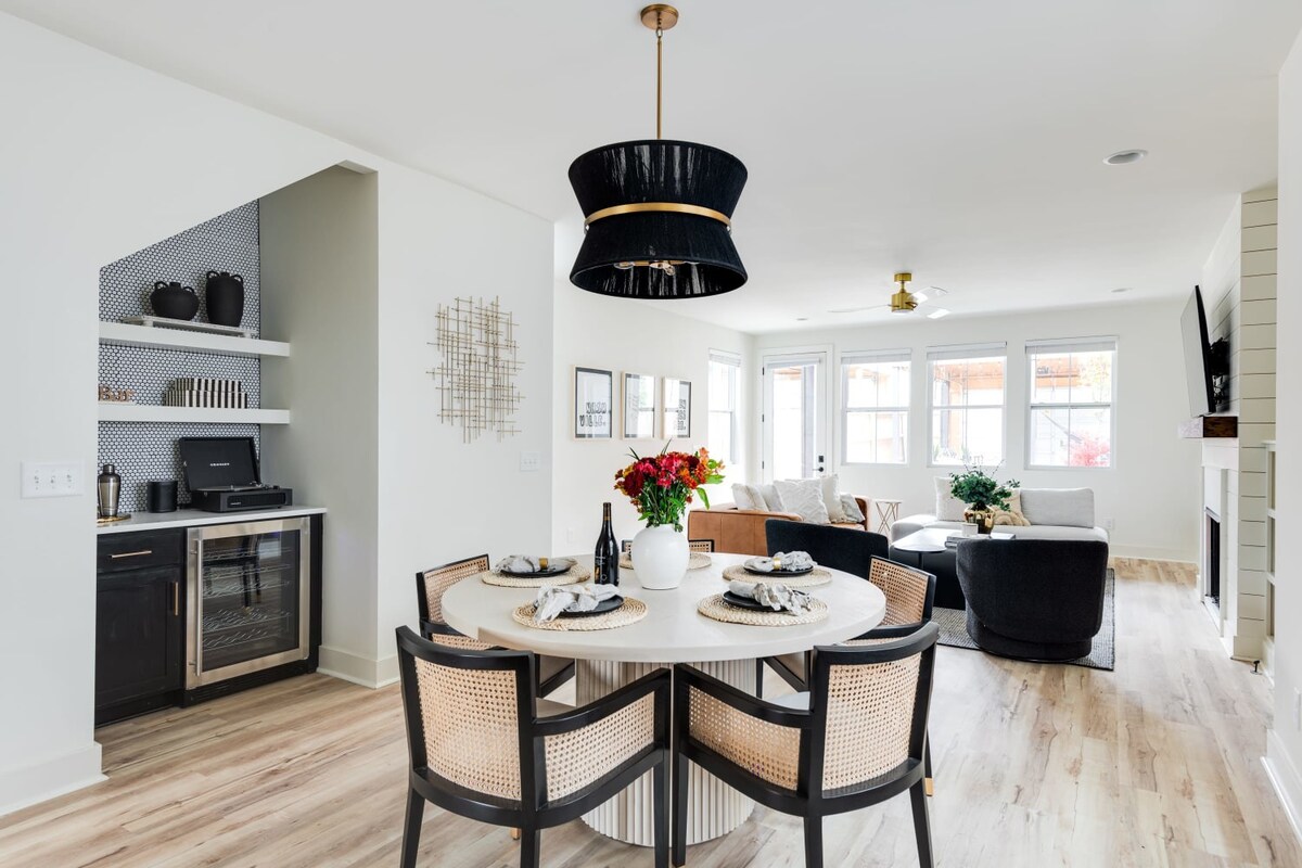 Elegant dining area with a round white table and four cane-back chairs, illuminated by a large black and gold chandelier. A built-in wet bar with a wine fridge, record player, and open shelving is tucked into a corner.
