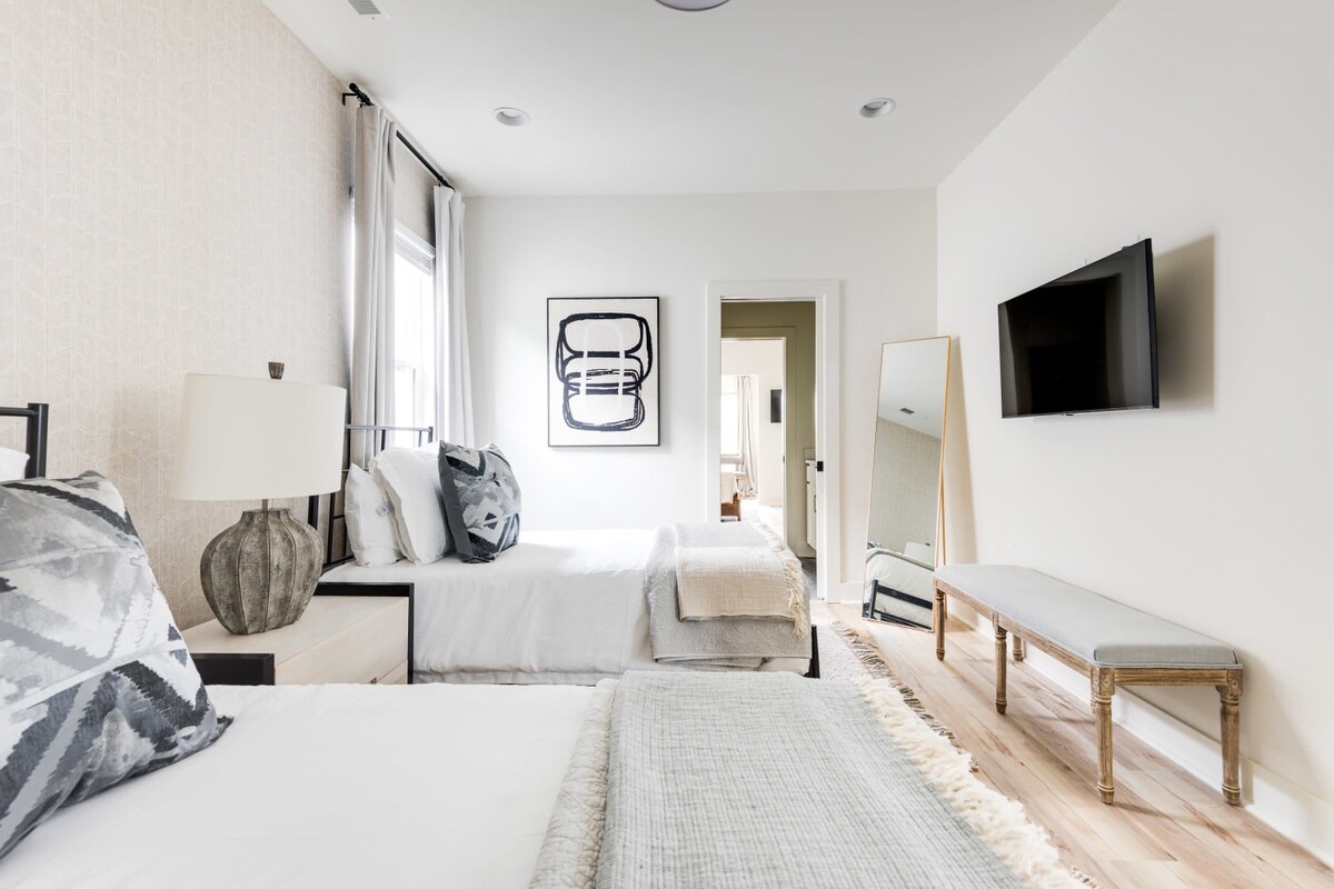 A close-up view of a bright bedroom showcasing two twin beds with white bedding and gray patterned pillows, flanking a white nightstand with a large gray lamp. A wall-mounted TV, full-length mirror, and upholstered bench are visible on the opposite wall.