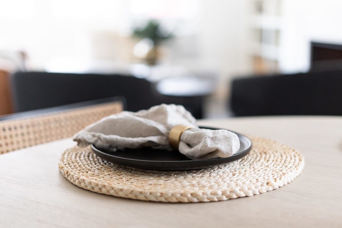 Close-up of a dining table place setting with a woven placemat, a black plate, and a linen napkin with a gold ring, showing a blurred background of the dining and living area.