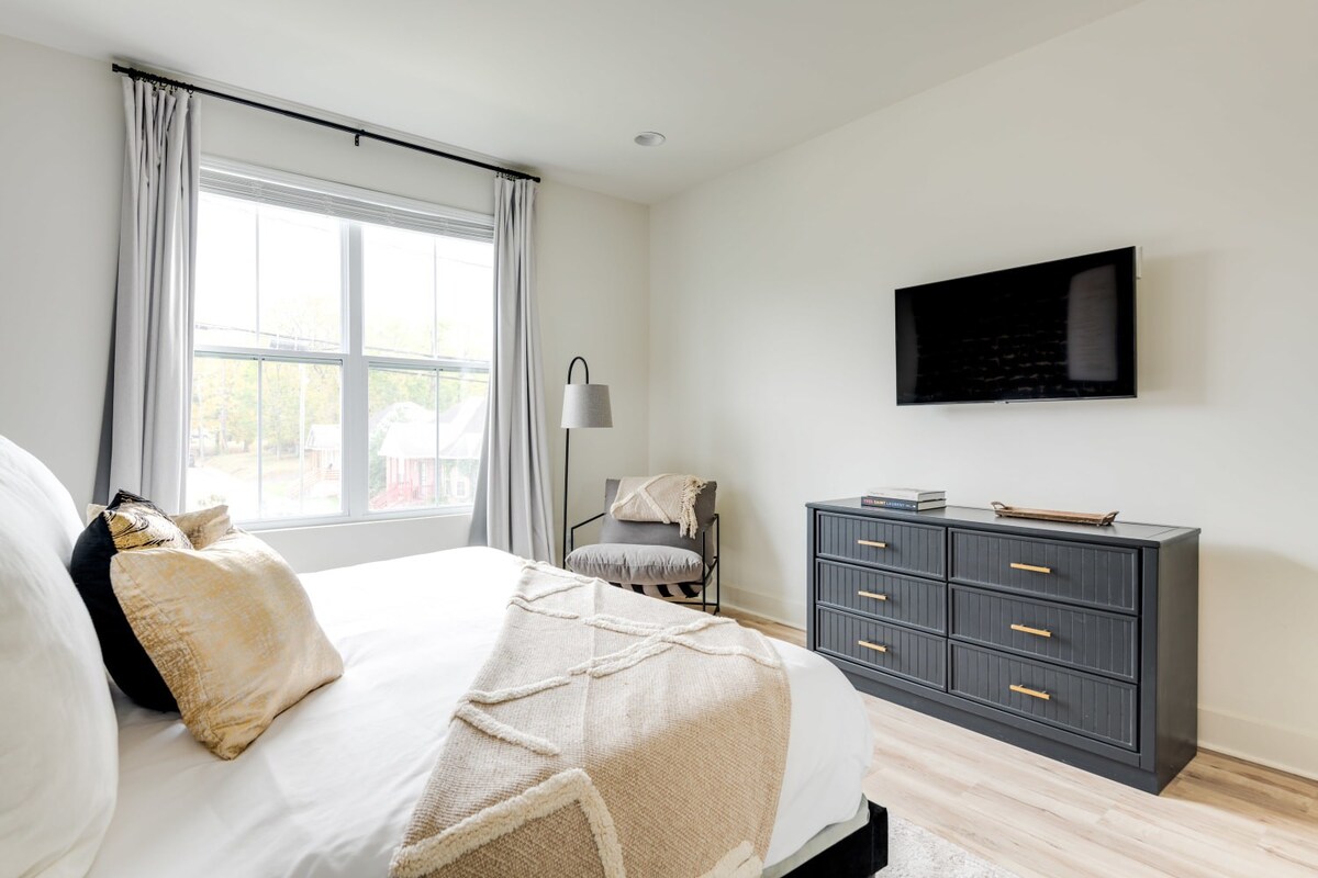 Bright bedroom with a bed dressed in white and beige, facing a large window with light grey curtains. A dark grey dresser with gold pulls sits beneath a wall-mounted TV, with a cozy armchair and floor lamp in the corner.