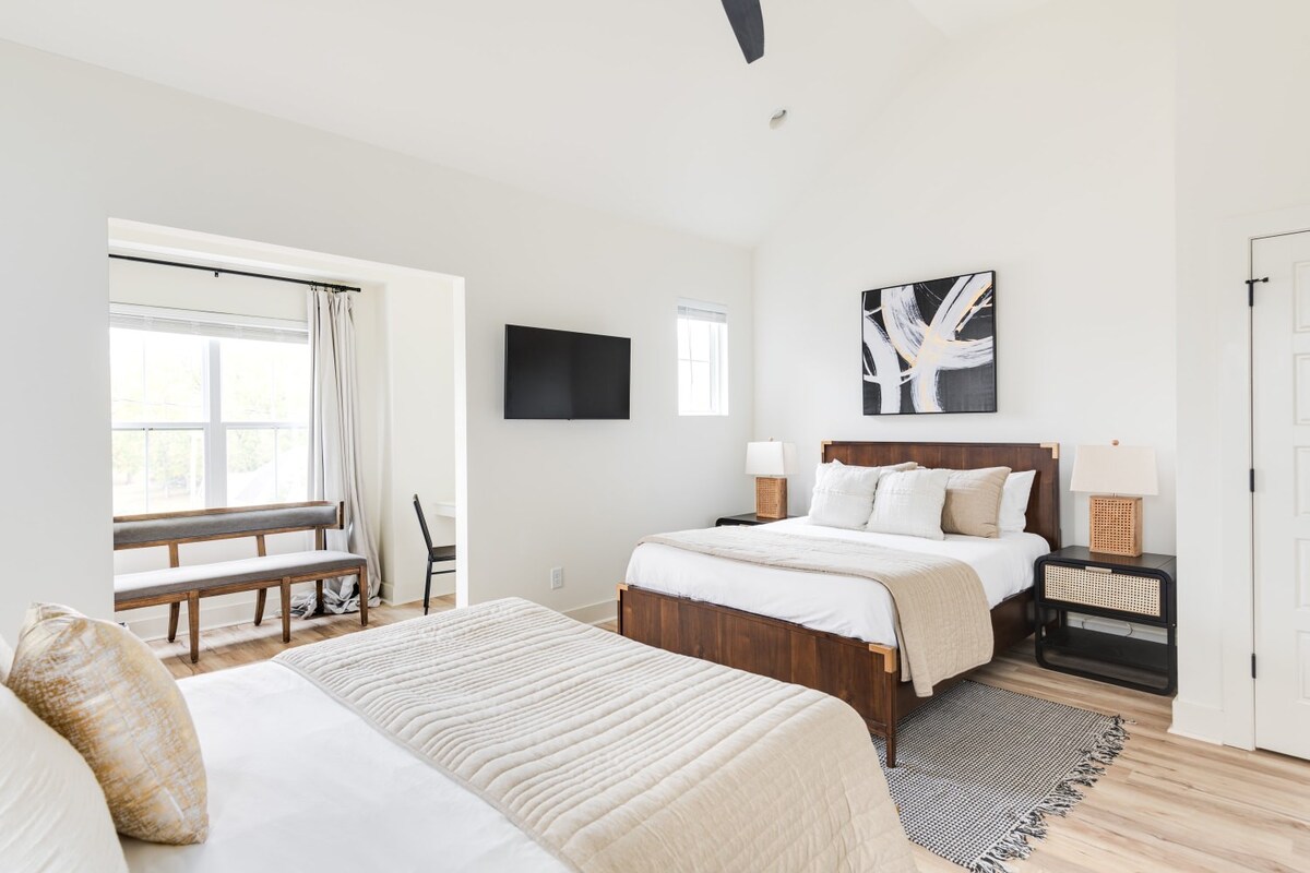 Expansive bedroom featuring two wooden beds with white and beige bedding, separated by a patterned rug. A second abstract art piece hangs above the far bed, and a windowed alcove with a wooden bench is visible in the background.
