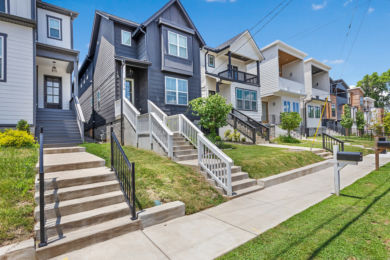 A row of modern houses with varying facades, including a prominent black house with white trim and multiple white staircases leading up from the sidewalk, set against a sunny sky.