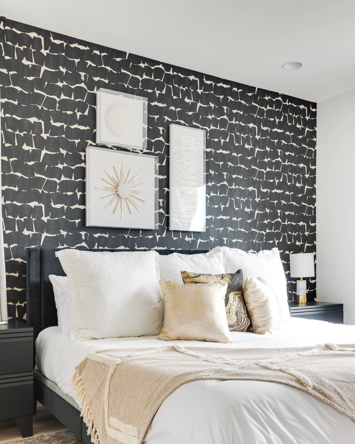A stylish bedroom featuring a bed with a dark upholstered headboard, white bedding, and gold accent pillows. A bold black and white patterned wallpaper creates a focal point behind the bed, complemented by three framed art pieces.