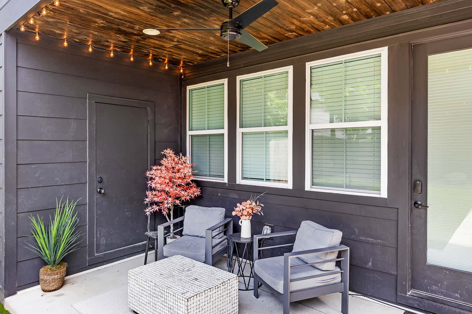 A cozy covered patio area with dark horizontal siding, furnished with two gray armchairs, a small round side table, and a white textured coffee table. The space is illuminated by string lights and features a ceiling fan.