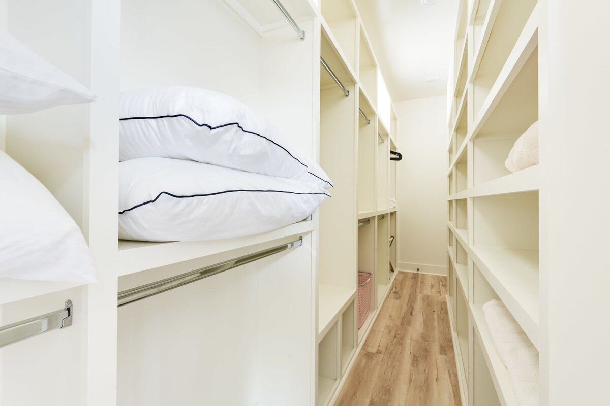 A spacious, well-organized walk-in closet with light wood flooring and white built-in shelving units on both sides, featuring hanging rods and neatly stacked white pillows.