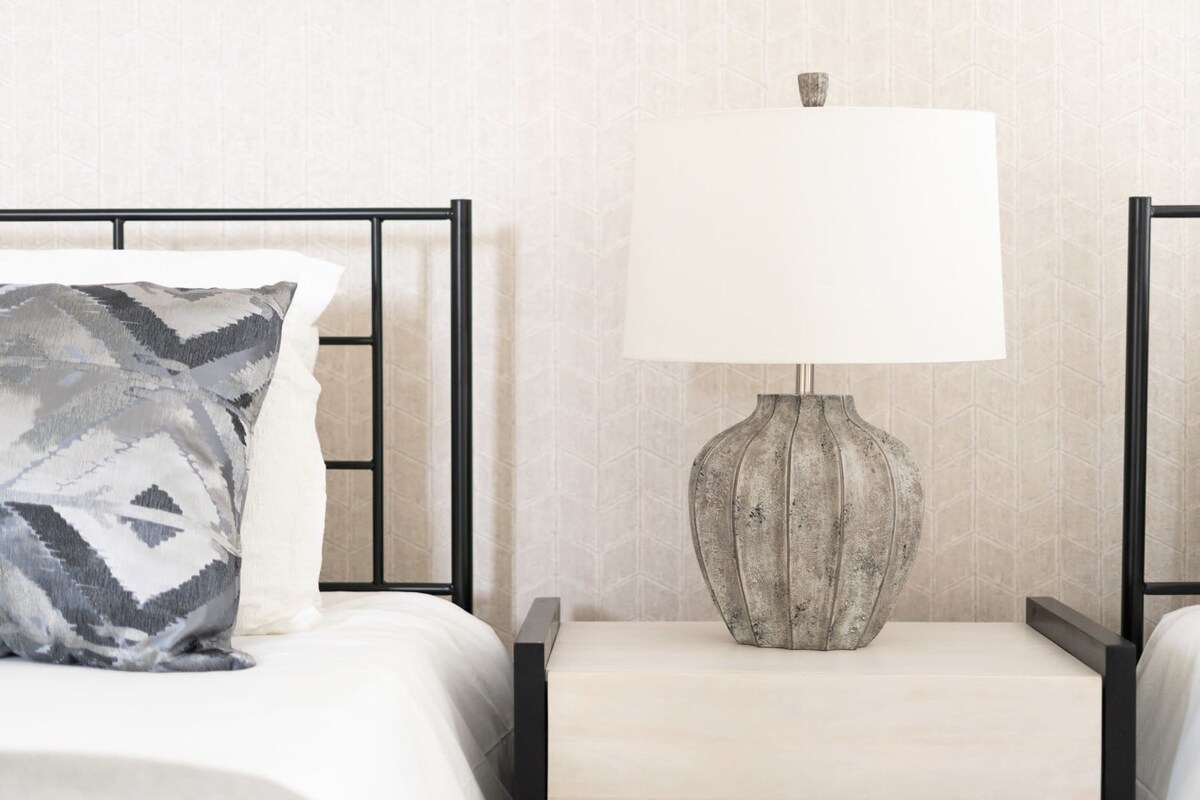 Close-up of a bedside area featuring a light-colored nightstand and a textured grey table lamp with a white shade. A black metal bed frame and subtle patterned wallpaper are visible in the background, creating a calm ambiance.