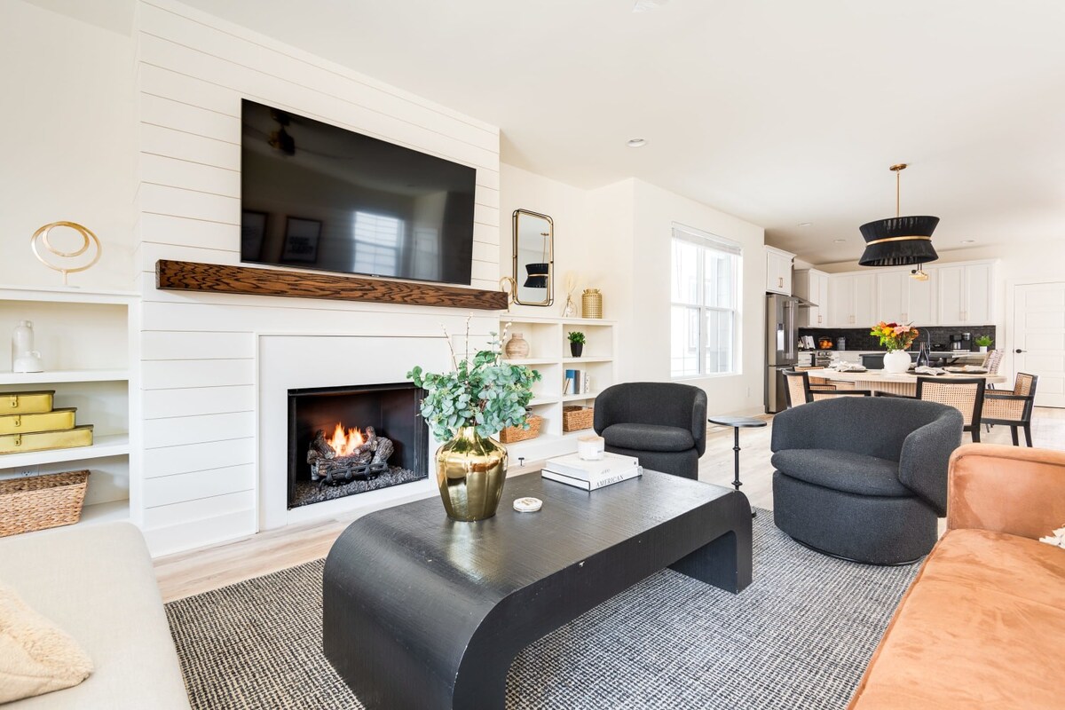 Angled view of the living room featuring a black coffee table with a gold vase, a fireplace with a TV above, white built-in shelves, and a glimpse of the dining area and kitchen in the background.