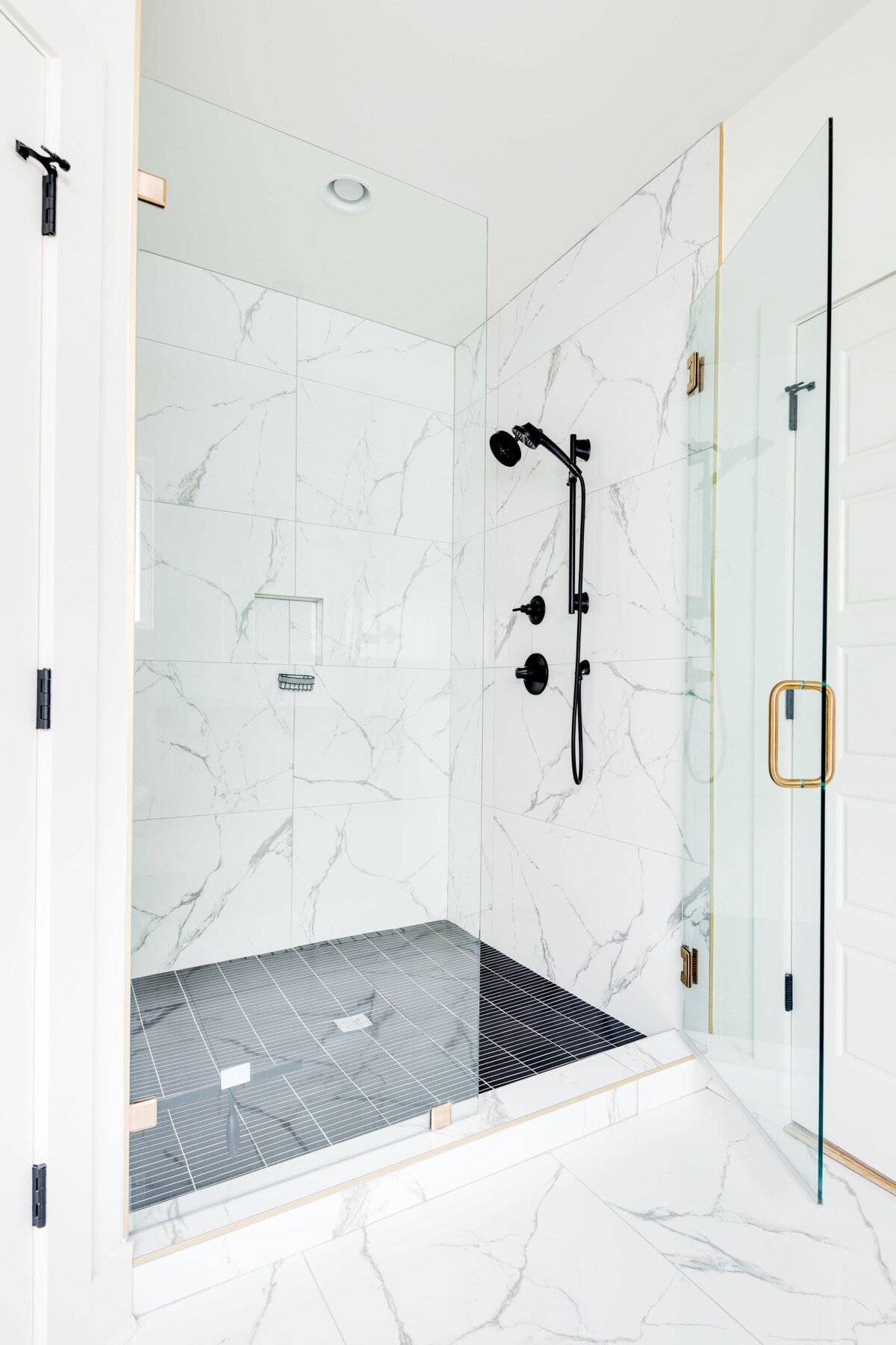 A modern walk-in shower with clear glass panels and black hardware, featuring white marble-look wall tiles and a dark gray tiled floor. A black rainfall showerhead and handheld sprayer are installed, along with a built-in niche.