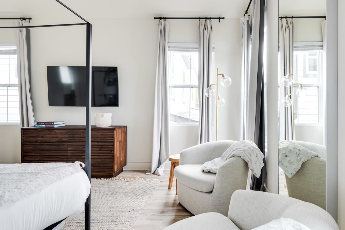 A bright bedroom featuring a black metal canopy bed frame, a wooden dresser with a mounted TV, and a cozy light gray armchair with a wooden side table. Light gray curtains frame the windows, and light wood flooring is covered by a textured rug.