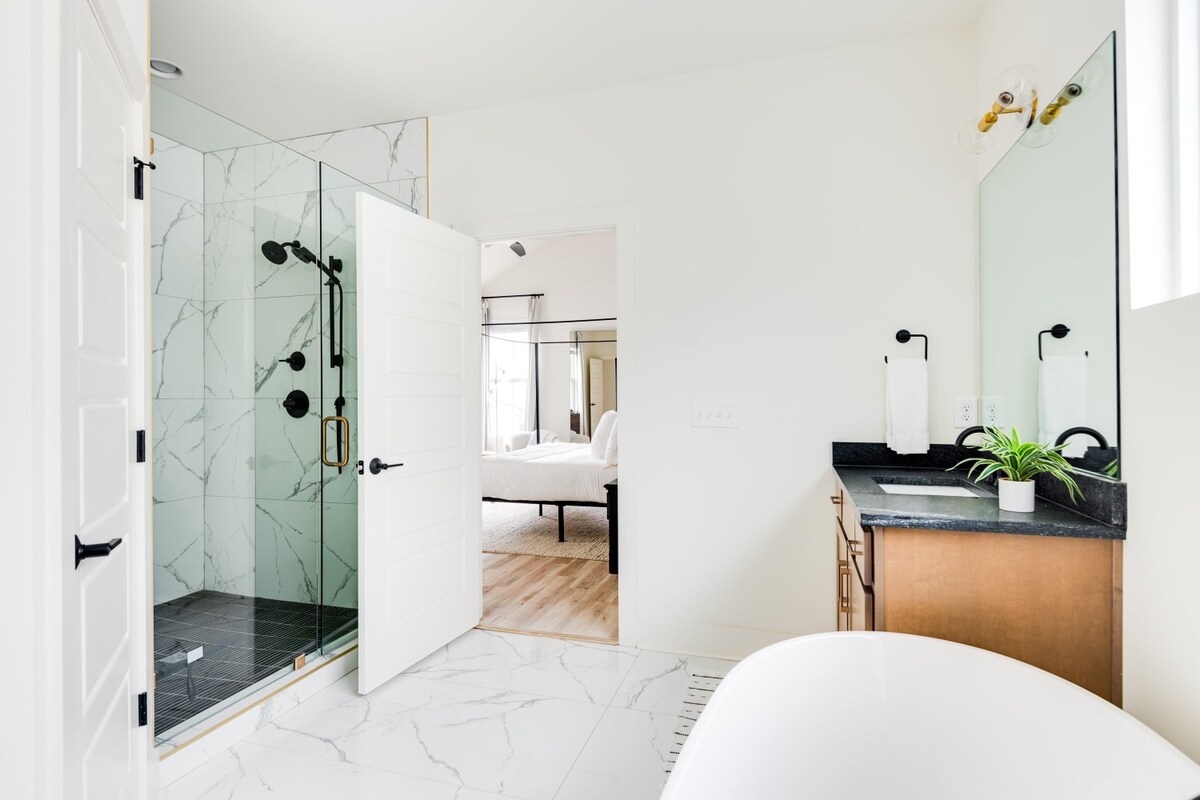 A bright bathroom with white marble-look floor tiles, featuring a glass-enclosed walk-in shower with black fixtures and white tiled walls. A light wood vanity with a dark countertop and mirror is visible, and an open door leads into a bedroom.