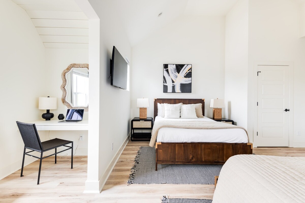 Serene bedroom with a dark wooden bed frame, white bedding, and light wood floors. An adjacent alcove features a white desk, black chair, and a decorative mirror, creating a functional workspace.