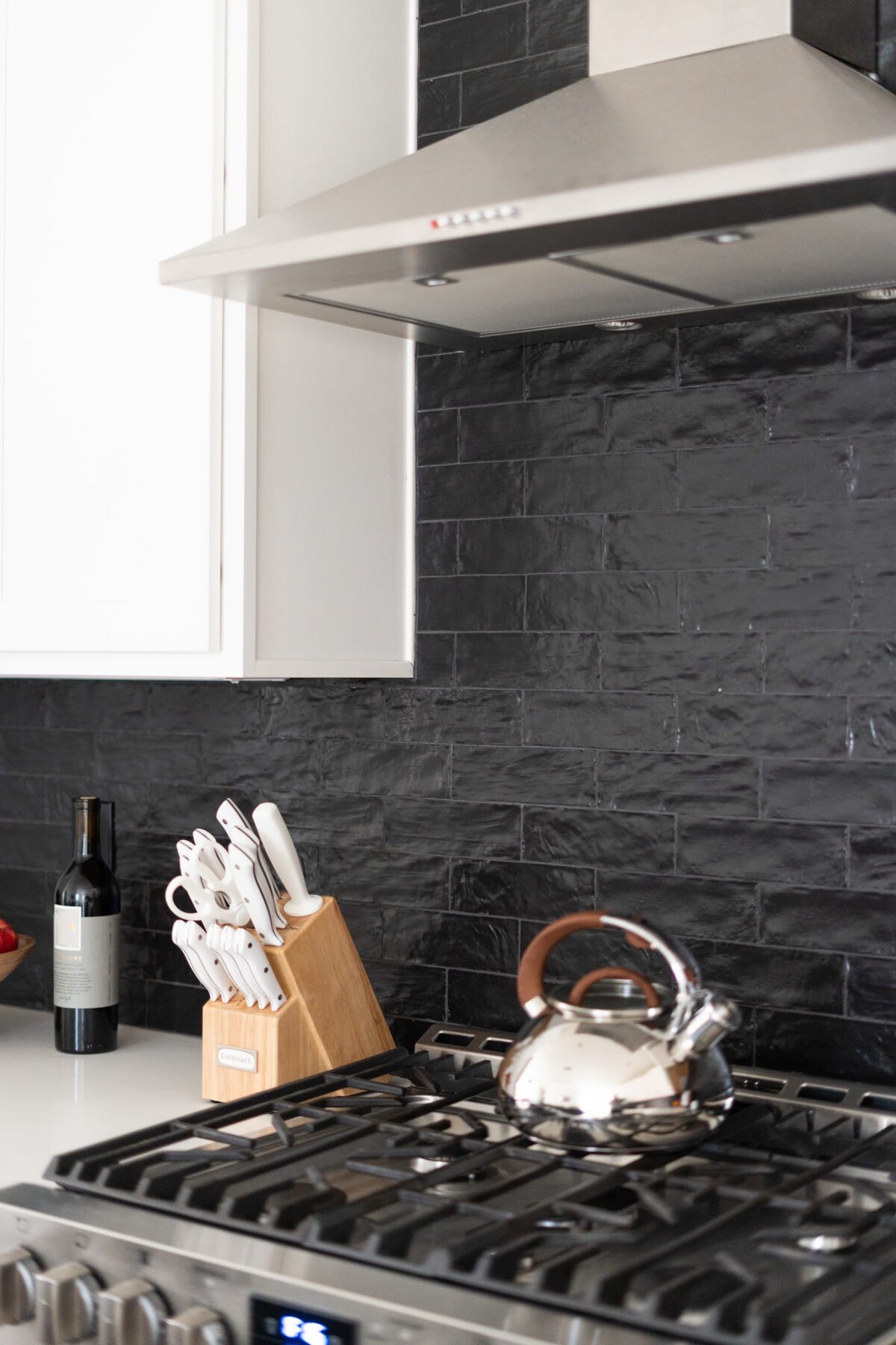 Close-up of a modern kitchen with a stainless steel range hood, gas stovetop, and a black textured subway tile backsplash, featuring a wooden knife block and a silver kettle.