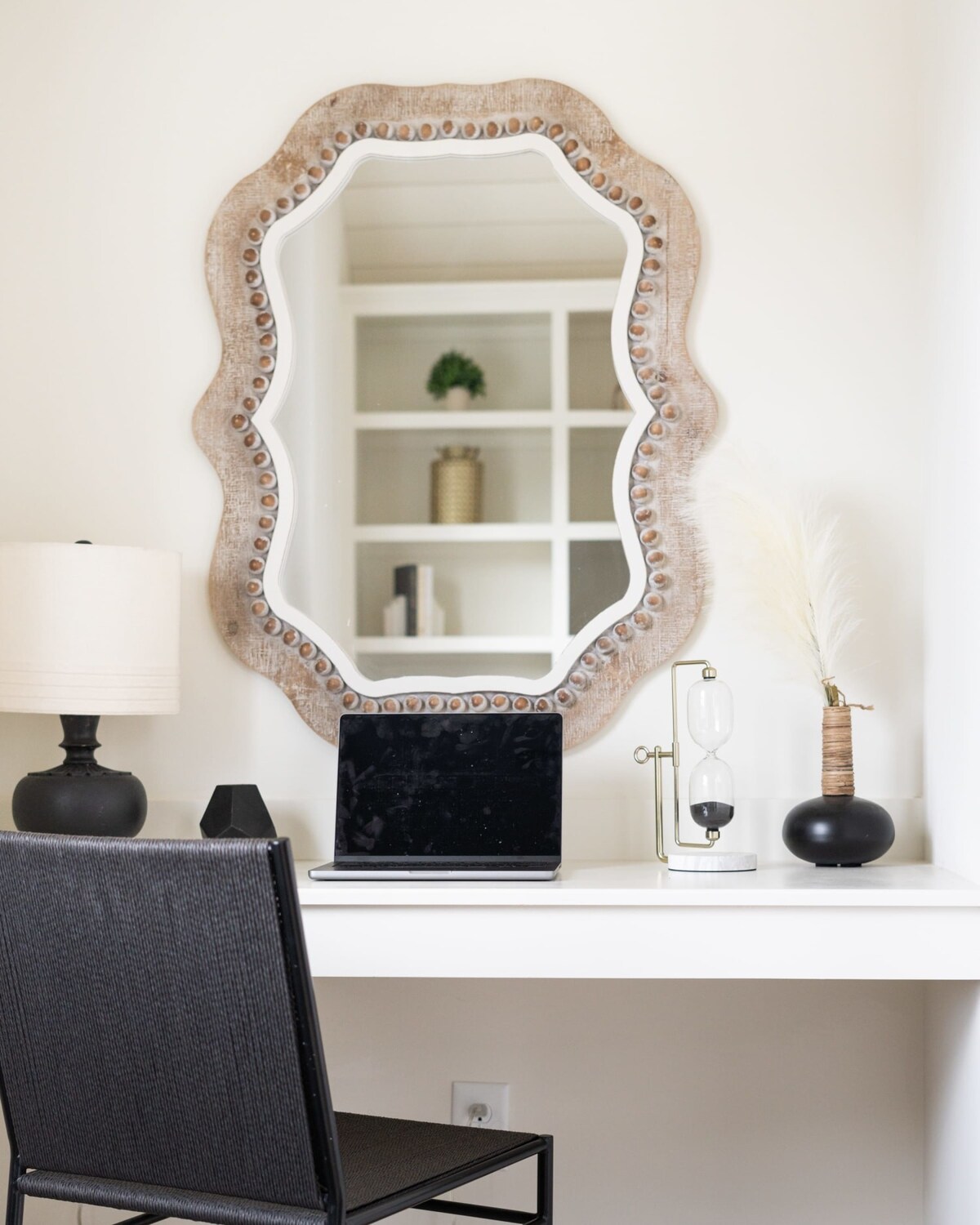 A dedicated workspace nook featuring a white desk with a laptop, a unique scalloped wooden mirror, and a black table lamp. Decorative hourglass and vase add a touch of elegance to the functional area.