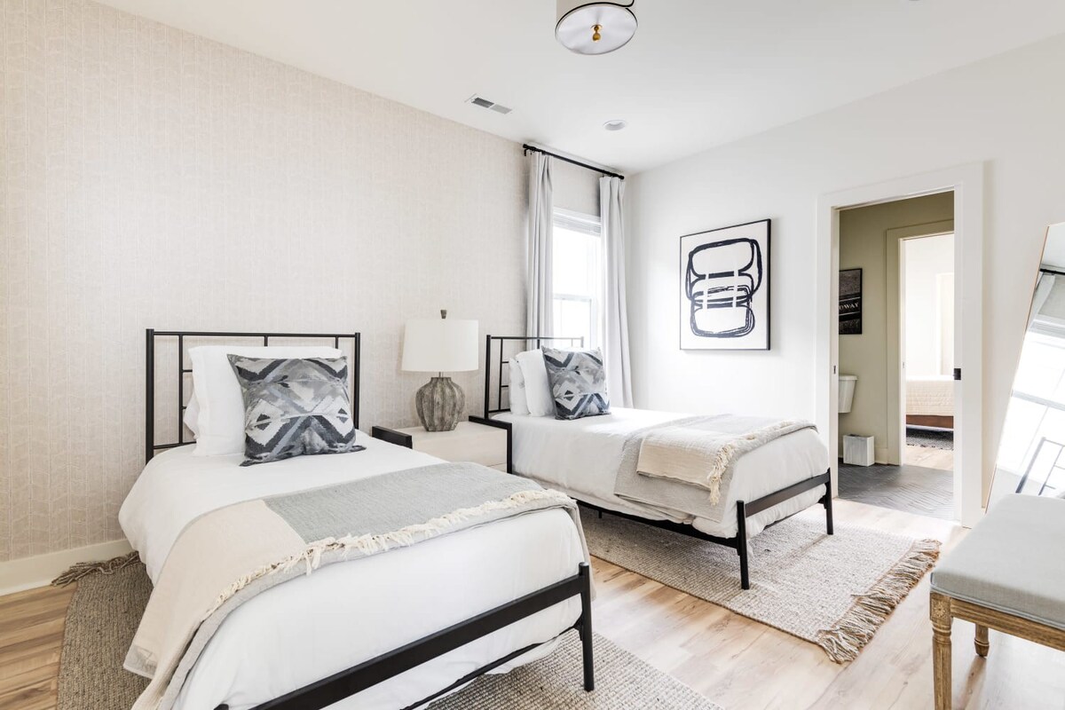 A serene bedroom with two twin beds, each featuring black metal frames, white bedding, and gray patterned pillows, separated by a white nightstand with a textured gray lamp. The room has light wood floors, textured rugs, and a light patterned accent wall.