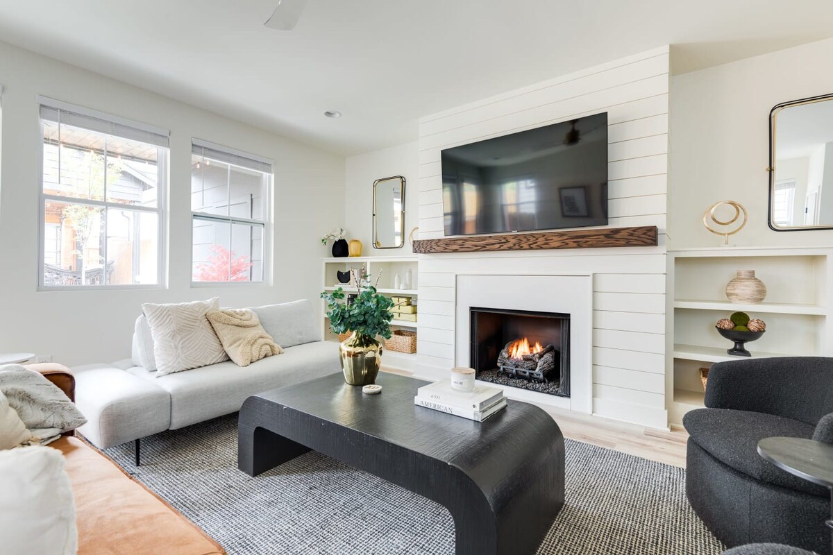 Cozy living room centered around a shiplap accent wall with a gas fireplace and mounted TV, flanked by built-in shelves. A light gray sectional and a brown leather sofa face each other across a dark, modern coffee table on a textured rug.