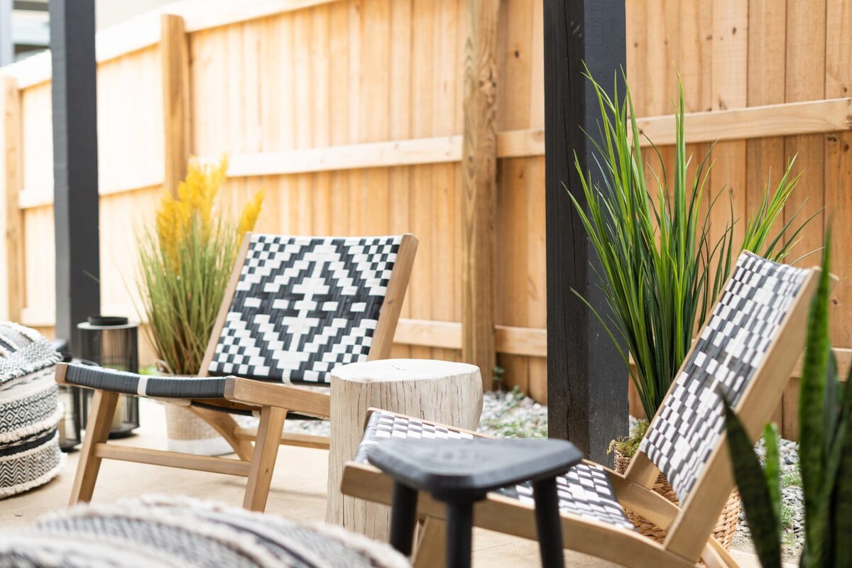 A close-up view of the outdoor patio shows two black and white patterned chairs, a wooden stump side table, and a patterned pouf, with tall green and yellow plants adding natural accents against a wooden fence. The concrete floor and covered ceiling are visible.