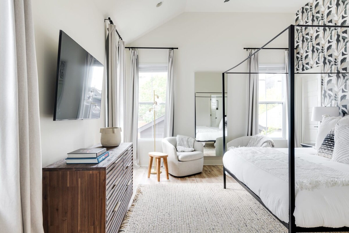 View of a bright bedroom with a black metal canopy bed, a wooden dresser with a wall-mounted TV, and a cozy armchair next to a large mirror, all on light wood-look flooring.