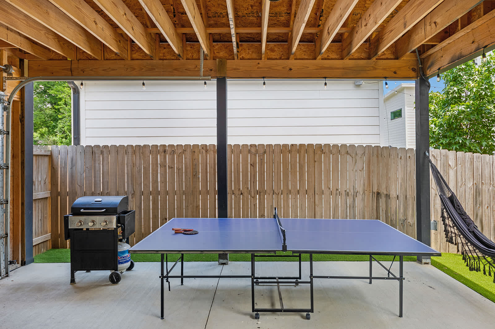 A covered outdoor recreation area with a wooden ceiling, featuring a blue ping-pong table, a gas grill, and a black hammock. The area has a concrete floor and is adjacent to an artificial turf section of the yard.