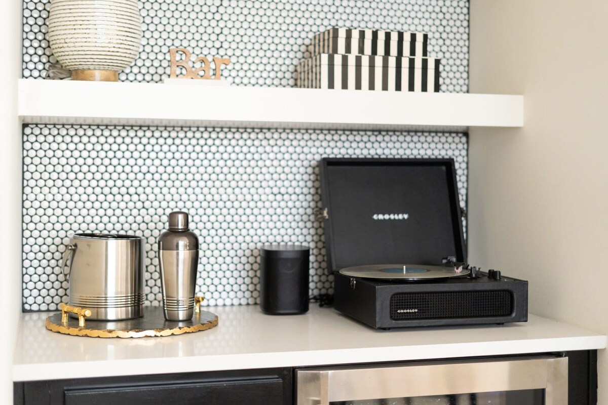 Close-up of a wet bar countertop with a silver ice bucket and cocktail shaker, a black speaker, and a black Crosley record player, all set against a black and white penny tile backsplash.