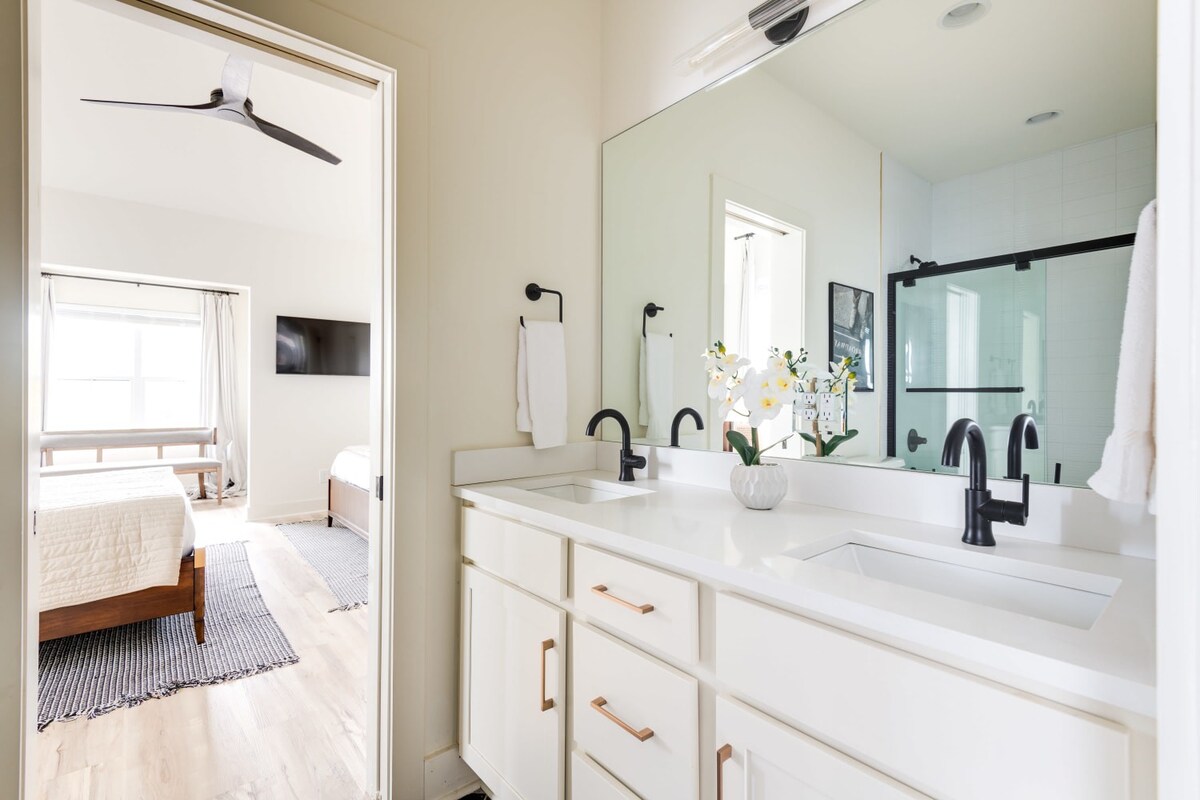 A bright bathroom featuring a double vanity with white quartz countertops, sleek black faucets, and white shaker-style cabinets with modern copper pulls. A large mirror reflects a bedroom with a bed and wall-mounted TV.