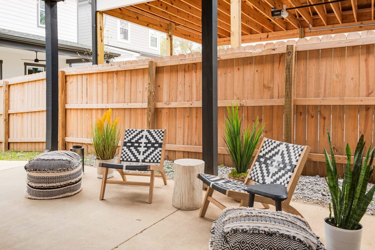 A cozy outdoor patio features two black and white patterned chairs, a wooden stump side table, and two large patterned poufs on a concrete slab, surrounded by a wooden fence and lush green plants. The area is covered by a wooden ceiling structure.