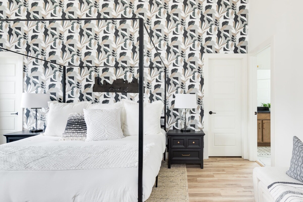 A wide view of the stylish bedroom with a black metal canopy bed, bold abstract leaf-patterned wallpaper, and black nightstands. Light wood flooring extends into the room, and a white paneled door is visible on the right, leading to another area.