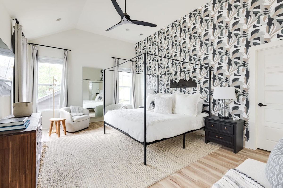 Spacious bedroom with a black metal canopy bed, a striking abstract wallpaper accent wall, light wood-look flooring, and a black ceiling fan.