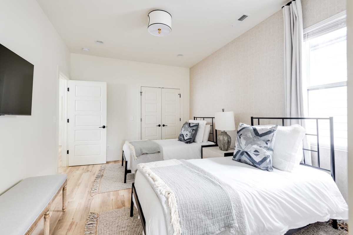 A bright bedroom featuring two twin beds with black metal frames, crisp white bedding, and geometric gray pillows, set against a light patterned accent wall and light wood flooring. A wall-mounted TV and a light gray upholstered bench are visible.