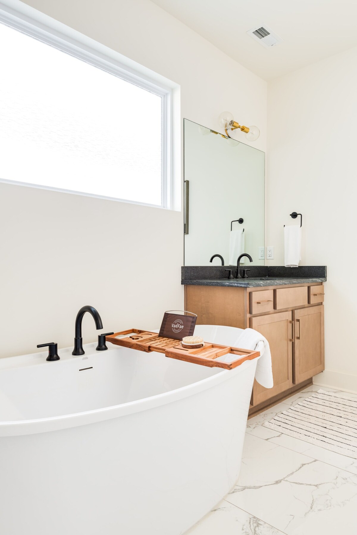 A close-up of the white freestanding soaking tub with black fixtures and a wooden bath caddy, set against a light wood vanity with a dark countertop and rectangular mirror. A horizontal window is positioned above the tub, providing natural light.