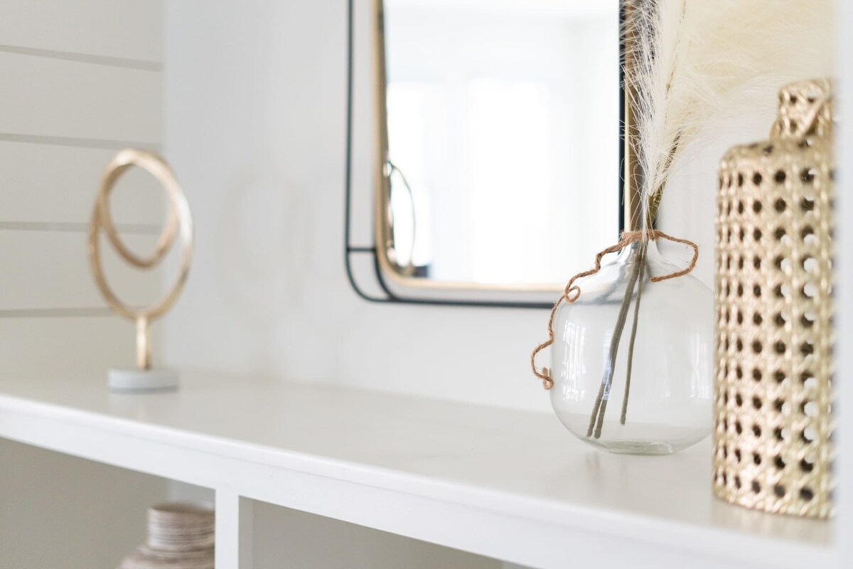 Detailed shot of the top shelf of a white built-in bookshelf, showcasing a gold abstract sculpture, a clear glass vase with pampas grass, and a gold cane-patterned vase, reflecting a modern aesthetic.