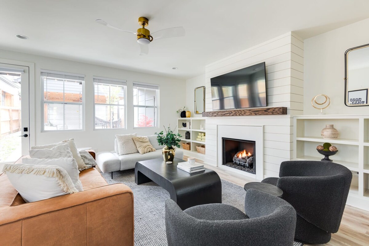 Bright living room featuring a shiplap accent wall with a mounted TV above a gas fireplace, flanked by built-in shelving. A light gray sectional and a brown leather sofa are arranged around a dark, modern coffee table on a patterned rug.