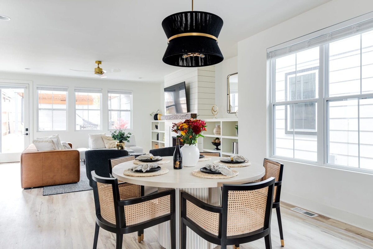 Bright open-concept dining area with a round light wood table and black and cane chairs, illuminated by a large black drum pendant light, leading into a living room with large windows.