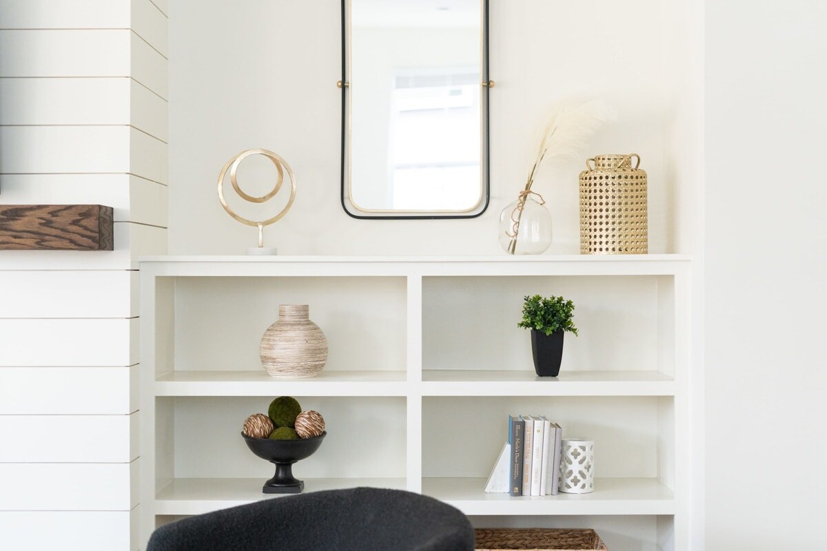 Close-up of a white built-in bookshelf adorned with decorative items including a gold abstract sculpture, a clear vase with pampas grass, a gold cane-patterned vase, and various books and bowls.