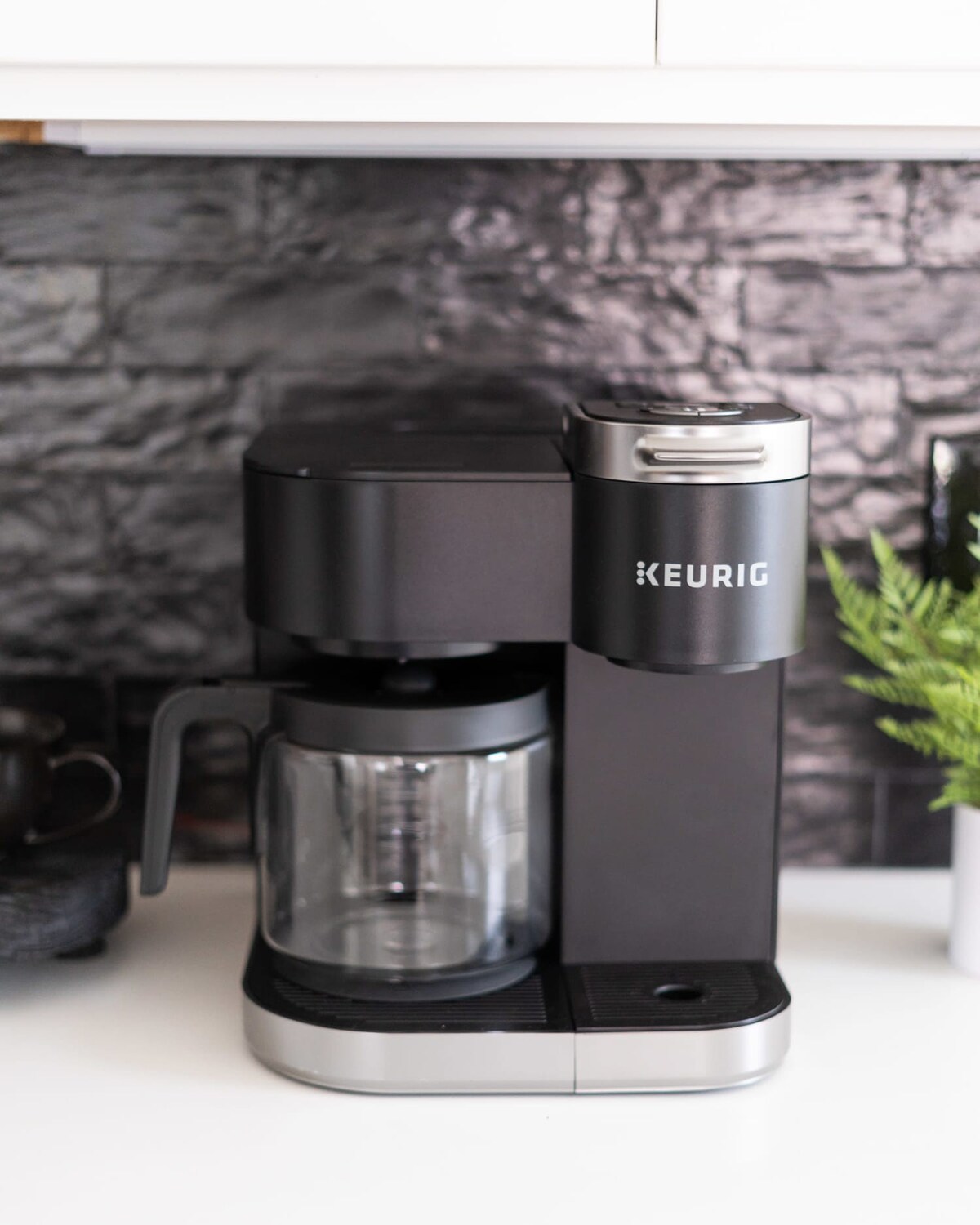 Close-up of a black Keurig coffee maker with a glass carafe on a white countertop, set against a black textured subway tile backsplash. A small green plant is visible to the right.
